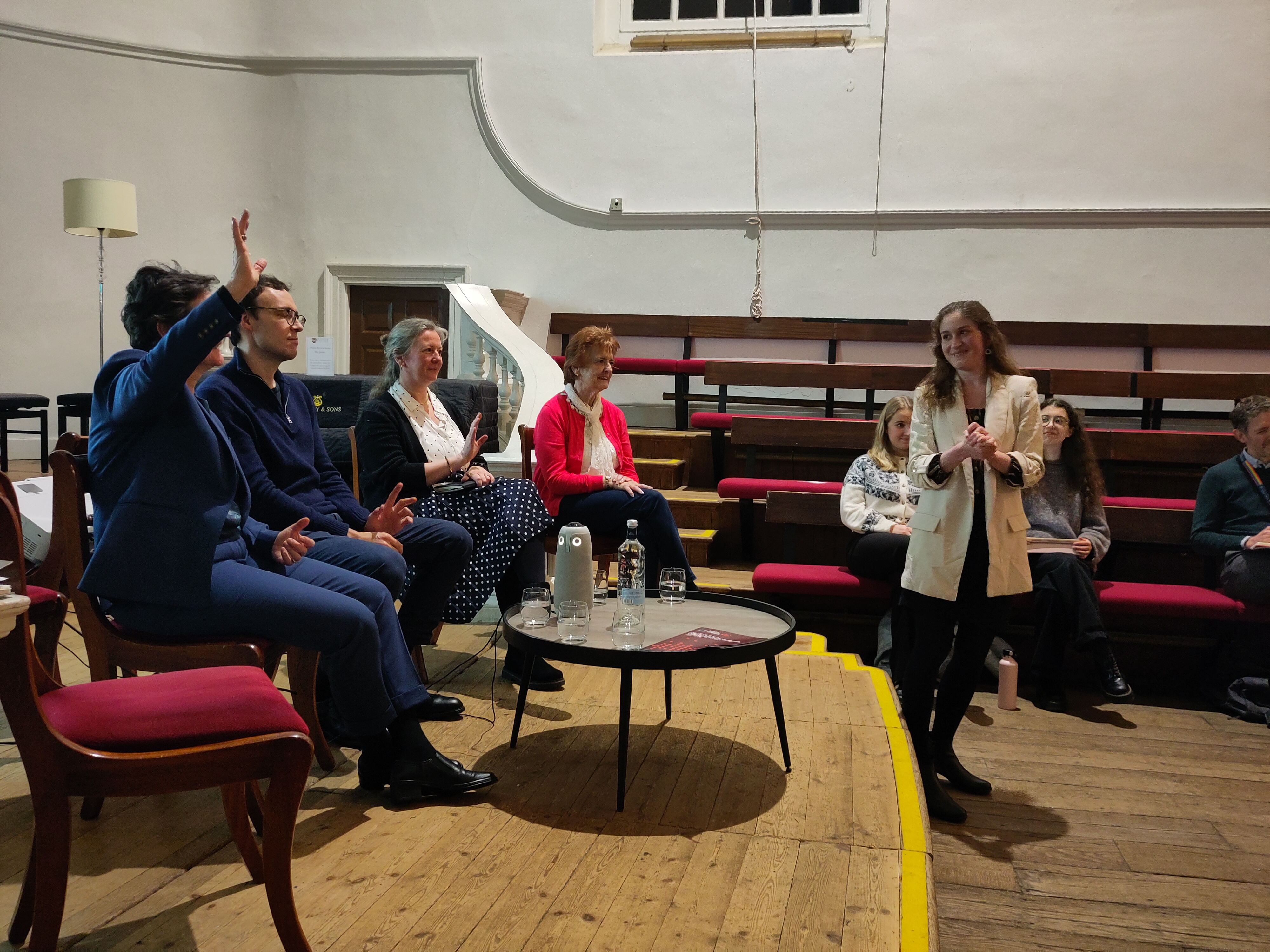 Luisa gestures back at the speakers, and the Vice Chancellor raises her hand.